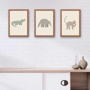 Puede incluir: Tres impresiones artísticas enmarcadas de animales. Las impresiones presentan un cocodrilo, un oso hormiguero y un mono en un estilo minimalista. Los animales están en tonos verdes y grises apagados sobre un fondo crema, enmarcados en madera. La obra de arte se exhibe en una pared de ladrillo blanco.