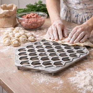 May include: A metal dumpling maker with a honeycomb design sits on a wooden surface. Ingredients like ground meat, flour, and dumplings are also visible. A person is using the dumpling maker to create dumplings.