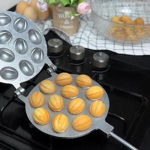 Könnte beinhalten: Ein silberner Metall-Walnuss-förmiger Keksmacher mit gebackenen Keksen. Die Kekse sind goldbraun und liegen auf der offenen Seite des Herstellers. Der Hersteller steht auf einem schwarzen Kochfeld. Die andere Seite des Herstellers ist offen.