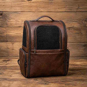 May include: A brown pet carrier backpack featuring a structured design with black mesh ventilation panels on the front and sides, a top handle, and multiple zippered compartments with dark metal hardware for secure storage and airflow.