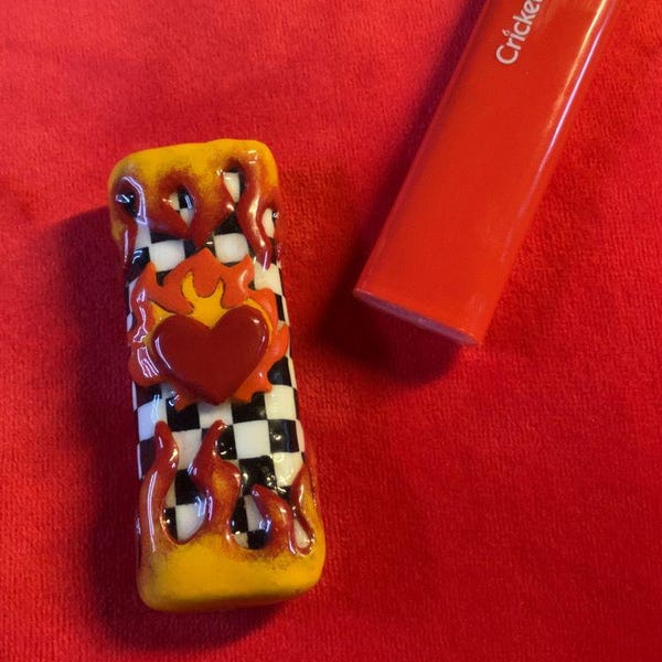 May include: A decorative lighter with a custom design and a red Cricket lighter. The custom design features a checkered pattern, a red heart, and flames. The lighter is on a red background.