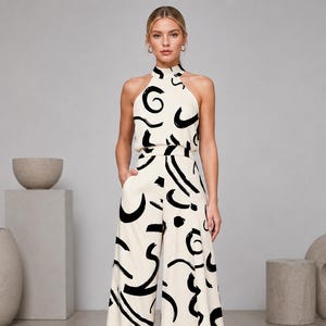 May include: A sleeveless jumpsuit in cream fabric featuring a bold black abstract brushstroke pattern. The garment has a high halter neckline, a fitted waist, and wide-leg trousers with side pockets, creating a continuous silhouette.