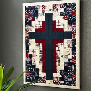 May include: A rectangular wall hanging quilt featuring a central dark navy blue cross shape. The background is composed of a patchwork of small fabric squares in red, white, and navy blue with various patterns including stars, stripes, and plaids.