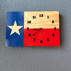 May include: A wooden wall clock featuring the Texas state flag design. The clock face is divided into blue, white, and red sections, with black numerals and hands. The clock measures approximately 12 inches by 10 inches.