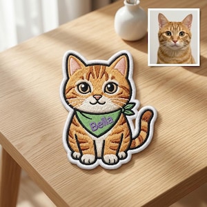 Könnte beinhalten: Gestickter Aufnäher mit einer orangefarbenen Tabby-Katze mit grünem Halstuch. Die Katze hat große Augen und der Name "Bella" ist auf dem Halstuch gestickt. Im Hintergrund ein gerahmtes Foto einer ähnlichen Katze.