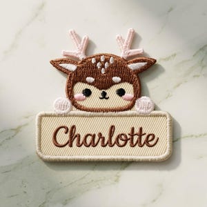 Op de afbeelding: Geborduurde patch met een cartoon hert met bruine vacht, roze geweien en een beige naamlabel. De naam "Charlotte" is in bruin schrift geborduurd. Het hert heeft zwarte ogen en roze wangen.