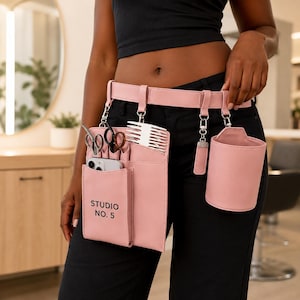 Peut inclure: Une ceinture utilitaire rose clair avec une pochette cylindrique détachable assortie et un organisateur rectangulaire portant l'inscription STUDIO NO. 5. L'organisateur contient des ciseaux argentés, un peigne blanc et un smartphone.