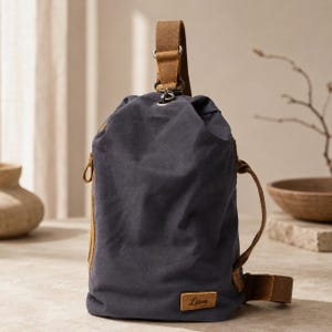 Puede incluir: Un bolso bandolera de lona gris oscuro con detalles en cuero marrón, una sola correa ajustable para el hombro y un asa lateral. El bolso tiene un parche de cuero rectangular en la parte inferior con el nombre Liam grabado en cursiva.