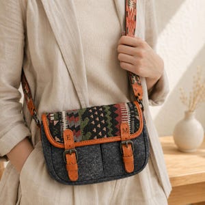 May include: A charcoal gray crossbody bag with a patterned flap featuring geometric shapes in red, green, and black. The bag has tan leather trim, two front buckle straps with the letters E and M, and a matching patterned adjustable shoulder strap.