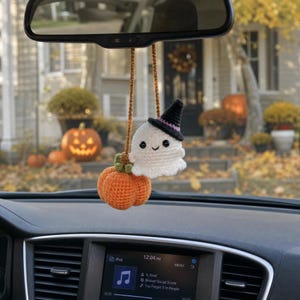 May include: A crocheted car accessory featuring a small white ghost wearing a black witch hat with a purple band, attached to a bright orange pumpkin with a green stem. The two figures are connected by a brown yarn cord for hanging.