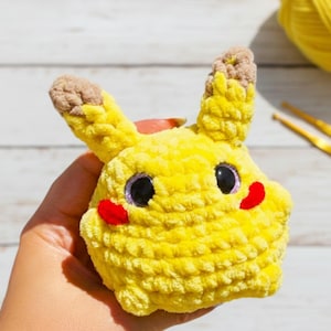 May include: A handmade, yellow crocheted Pikachu plushie with black eyes, red cheeks, and brown-tipped ears. The plushie is held in a hand, with a ball of yellow yarn and crochet hooks in the background. The plushie is a popular character from the Pokemon franchise.