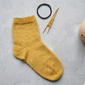 Może przedstawiać: Musztardowa dzianinowa skarpeta z prążkowanym mankietem, drutem okrągłym i drutami dwustronnymi. Skarpeta jest ręcznie robiona i wydaje się być wykonana z wełny lub podobnej przędzy. Przedmioty ułożone są na jasnoszarej powierzchni.