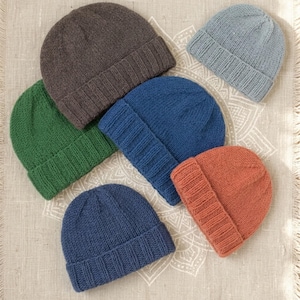 May include: A collection of six knitted beanies in various solid colors including dark brown, light blue, forest green, royal blue, and terracotta orange. Each beanie features a thick ribbed cuff and a smooth stockinette stitch crown.