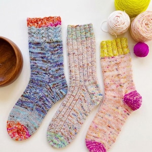 May include: Three hand-knitted socks featuring varied colour palettes. The left sock has blue and purple variegated yarn with an orange cuff and toe. The middle sock is light beige with multicoloured flecks. The right sock is peach with a lime cuff and magenta toe.
