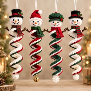 May include: Four crocheted snowman ornaments hanging vertically. Each features a white head with a carrot nose, black eyes, and a smile. They wear different hats, scarves, and have spiral-shaped bodies in red, white, and green yarn colors.
