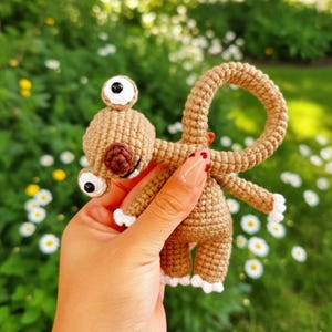 Può includere: Una creatura all'uncinetto fatta a mano nei toni del marrone, con due grandi occhi bordati di bianco e pupille nere. Il design stravagante include un anello e piccole braccia e gambe con dettagli bianchi. Il giocattolo è tenuto contro uno sfondo di fogliame verde e fiori bianchi.