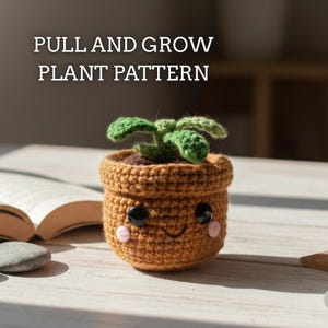 Puede incluir: Una maceta de ganchillo con una cara sonriente, ojos de botón negros y mejillas rosas. La maceta contiene una pequeña planta verde y está etiquetada como "PULL AND GROW PLANT PATTERN". La maceta es marrón y está sobre una superficie blanca.