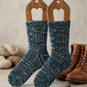 Puede incluir: Un par de calcetines de punto azul y gris exhibidos en hormas de madera. Los calcetines tienen un aspecto texturizado con una mezcla de hilos azules, grises y blancos. En el fondo se ven botas de cuero marrón, lo que sugiere un tema invernal y acogedor.