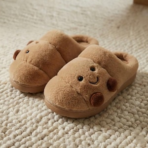 Op de afbeelding: Een paar bruine pluche pantoffels in de vorm van lachende stripfiguren. De pantoffels hebben een zachte, donzige textuur, met donkerbruine accenten voor de ogen, neus en mond. De zolen zijn donkerbruin.