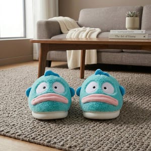 Op de afbeelding: Een paar novelty pantoffels in de vorm van vissen. De pantoffels zijn turquoise met witte ogen, roze lippen en blauwe vinnen. Ze hebben een pluche textuur en witte zolen.