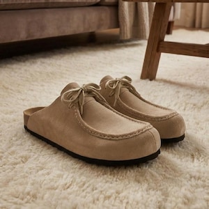 Puede incluir: Un par de zuecos de ante beige con cordones, colocados sobre una alfombra blanca y esponjosa. Los zuecos tienen una puntera redondeada y una costura cosida alrededor de la parte superior. Las suelas son negras y los zapatos parecen nuevos.