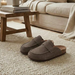 Peut inclure: Une paire de sabots en daim gris avec une semelle en liège, posée sur un tapis crème. Les sabots ont une bride sur le dessus et une semelle en caoutchouc marron. L'arrière-plan comprend un tabouret en bois, des livres et un canapé.