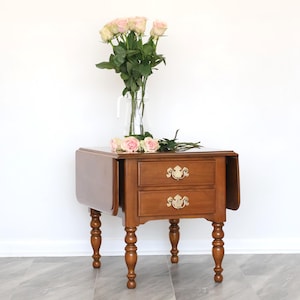 May include: A small wooden side table with two drawers and drop leaves. The table features decorative brass handles and turned legs. A glass vase holds pink roses, with more roses on the table. The table is a warm brown colour. The table is approximately 60 cm high.