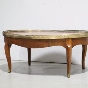 May include: An oval wooden coffee table featuring a smooth brown surface and a decorative brass gallery rail around the edge. The table is supported by four curved wooden legs, each accented with small brass feet at the bottom of the legs.
