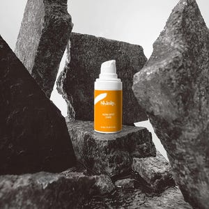 May include: A skincare product bottle with a white pump and an orange label, featuring the text "Skinity. ACNE SPOT CARE". The bottle is set against a backdrop of dark gray rocks, with water at the base.