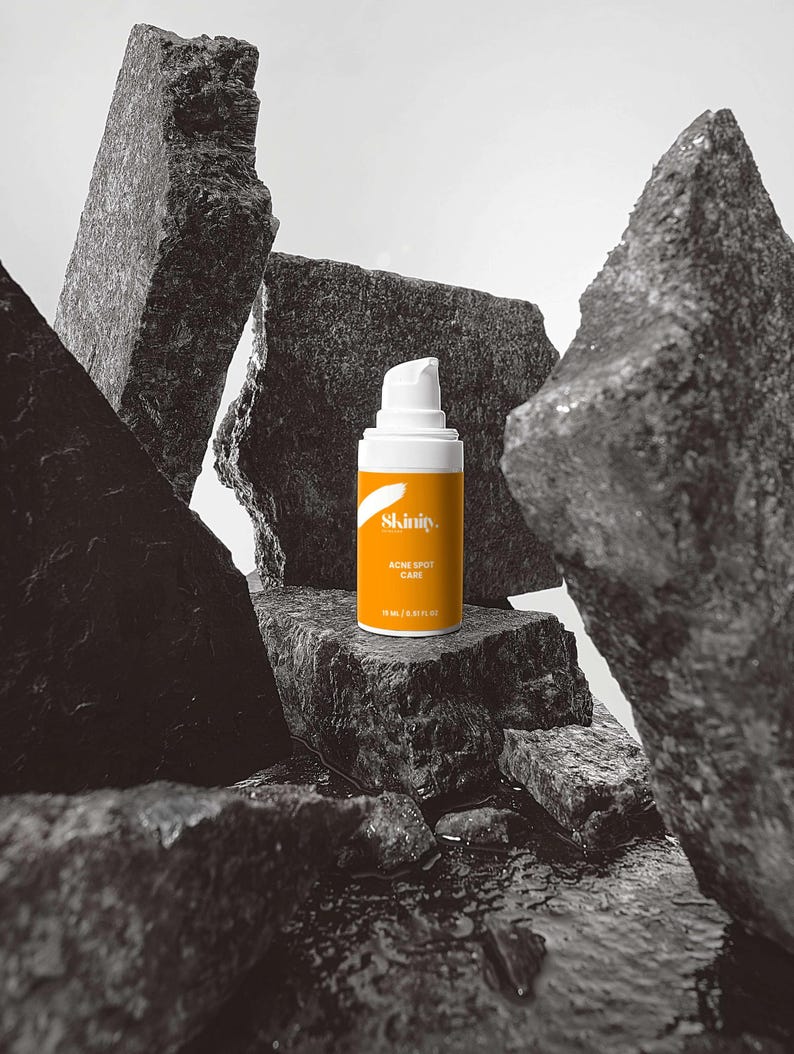 May include: A skincare product bottle with a white pump and an orange label, featuring the text "Skinity. ACNE SPOT CARE". The bottle is set against a backdrop of dark gray rocks, with water at the base.