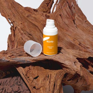 May include: A skincare product bottle with a white pump and a bright yellow label. The label reads "Skinity. Acne Spot Care" and the bottle is 15 ml / 0.51 fl oz. The bottle is next to its cap, and both are on a textured, brown wooden surface.