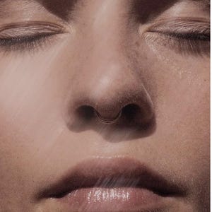 May include: Close-up of a person's face with closed eyes, a septum piercing, and full lips. The skin has a warm, natural tone, and the image is softly lit, creating a serene and intimate feel. The focus is on the facial features.