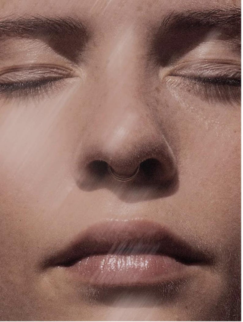 May include: Close-up of a person's face with closed eyes, a septum piercing, and full lips. The skin has a warm, natural tone, and the image is softly lit, creating a serene and intimate feel. The focus is on the facial features.