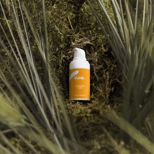 May include: A white and orange skincare product bottle with the text "Skinity. Acne Spot Care" is set against a backdrop of green moss and tall grass. The bottle has a pump dispenser and contains 15 ml / 0.51 FL OZ of product.