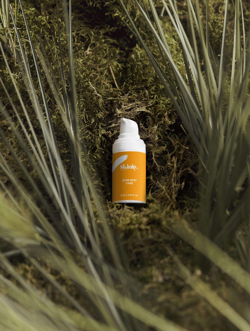 May include: A white and orange skincare product bottle with the text "Skinity. Acne Spot Care" is set against a backdrop of green moss and tall grass. The bottle has a pump dispenser and contains 15 ml / 0.51 FL OZ of product.