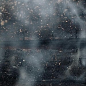 May include: Close-up of charred wood with wisps of smoke rising. The wood is dark brown and black, with visible cracks and textures. Small, light-colored particles are scattered across the surface, creating a contrast with the dark background. The image evokes a sense of heat and burning.