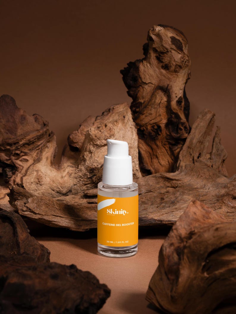 May include: A clear glass bottle with a white pump top and a bright yellow label. The label reads "Skinity Caffeine Gel Booster" and contains 30 mL / 1.01 fl oz. The bottle is set against a backdrop of brown wood.