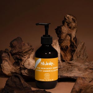 May include: A black bottle of Skinity Silk Skin Body Serum with a black pump dispenser and a yellow label. The label includes the text "WOODY AMBER & LEATHER" and "270 ML / 9.13 FL OZ". The bottle is set against a brown background with wood.