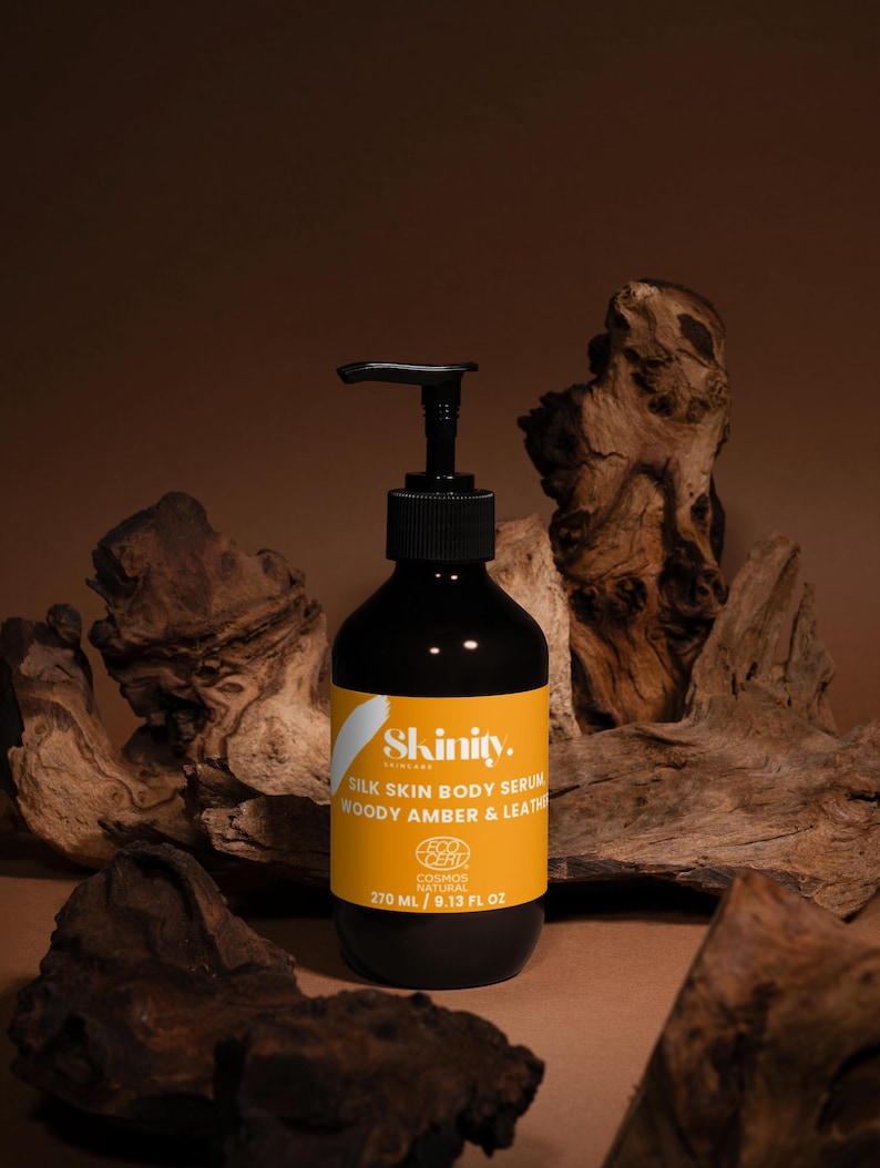 May include: A black bottle of Skinity Silk Skin Body Serum with a black pump dispenser and a yellow label. The label includes the text "WOODY AMBER & LEATHER" and "270 ML / 9.13 FL OZ". The bottle is set against a brown background with wood.