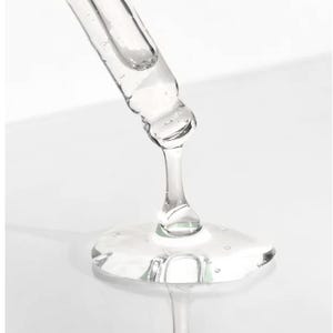 May include: A clear glass dropper dispensing a viscous, transparent liquid. The liquid forms a small, reflective puddle. The dropper is angled downward, with the tip close to the surface. The background is a soft, white gradient.