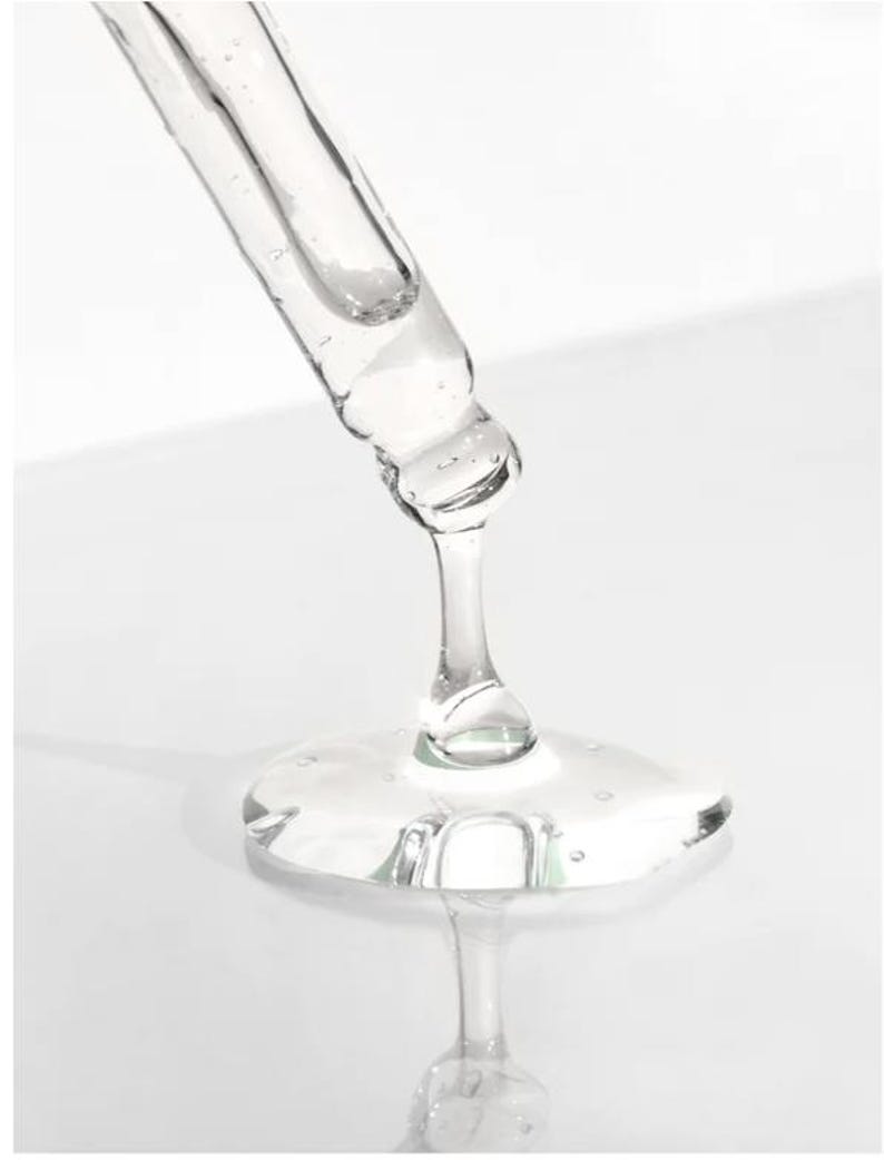 May include: A clear glass dropper dispensing a viscous, transparent liquid. The liquid forms a small, reflective puddle. The dropper is angled downward, with the tip close to the surface. The background is a soft, white gradient.