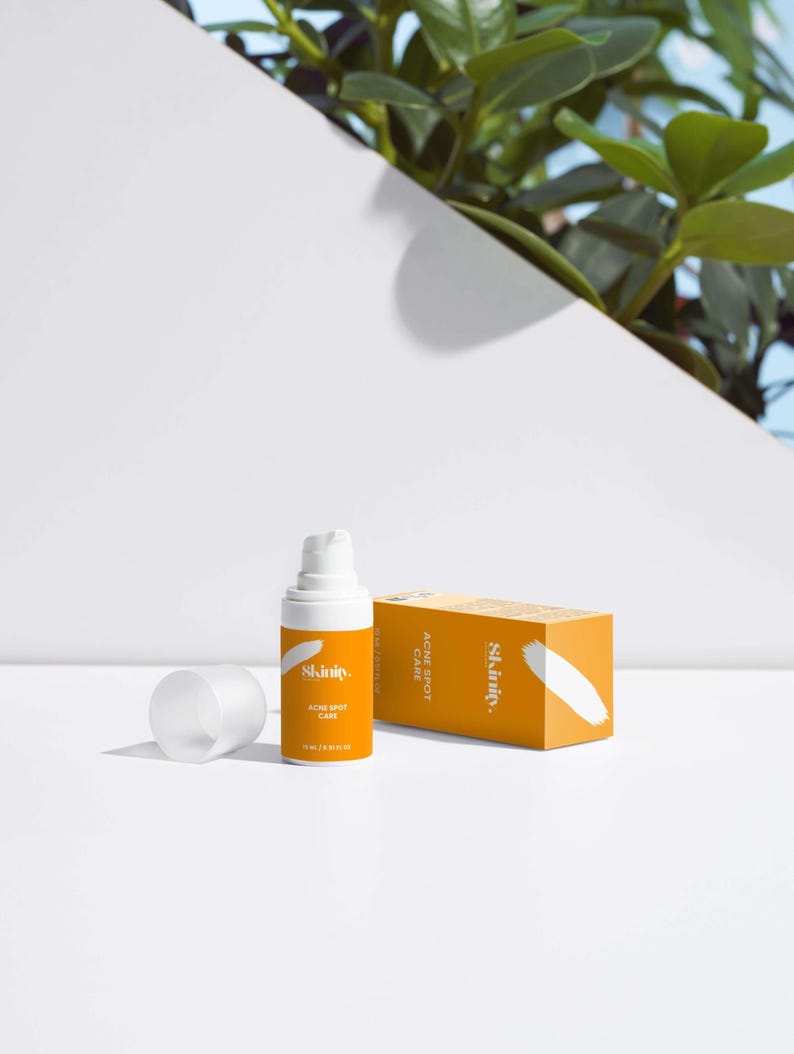 May include: An orange skincare product bottle and box with the brand name "Skinity" and the words "Acne Spot Care". The bottle is white with a pump dispenser. The box is rectangular and has a white brushstroke design.