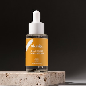 K&ouml;nnte beinhalten: Eine klare Glasflasche mit wei&szlig;er Pipette und gelbem Etikett mit der Aufschrift "Skinity AHA Peeling Concentrate". Die Flasche enth&auml;lt 30 ml Produkt und steht auf einer strukturierten, hellfarbigen Oberfl&auml;che vor dunklem Hintergrund.