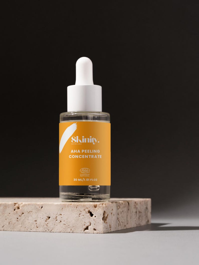 K&ouml;nnte beinhalten: Eine klare Glasflasche mit wei&szlig;er Pipette und gelbem Etikett mit der Aufschrift "Skinity AHA Peeling Concentrate". Die Flasche enth&auml;lt 30 ml Produkt und steht auf einer strukturierten, hellfarbigen Oberfl&auml;che vor dunklem Hintergrund.