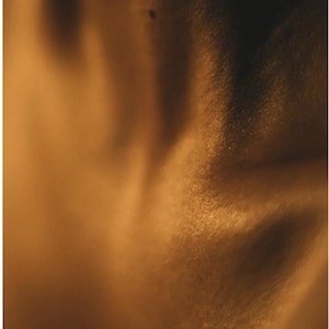 May include: A close-up shot of a person's skin, illuminated with warm, golden light. The skin texture is visible, with subtle shadows and highlights creating a soft, diffused effect. A small mole is visible.