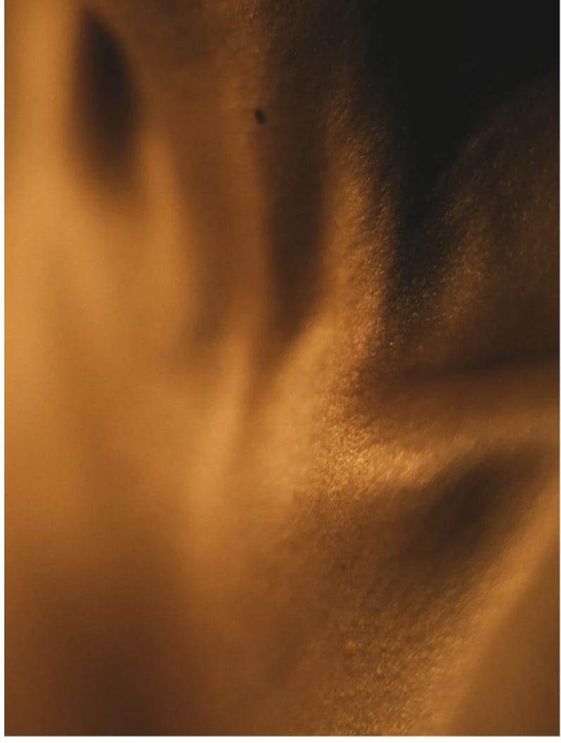 May include: A close-up shot of a person's skin, illuminated with warm, golden light. The skin texture is visible, with subtle shadows and highlights creating a soft, diffused effect. A small mole is visible.