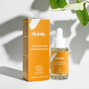 K&ouml;nnte beinhalten: Eine orangefarbene Schachtel und eine transparente Glasflasche mit Hautpflegeprodukten werden vor einem wei&szlig;en Hintergrund mit gr&uuml;nen Bl&auml;ttern gezeigt. Auf der Schachtel und der Flasche steht der Markenname "Skinity" und der Text "AHA Peeling Concentrate". Die Flasche enth&auml;lt 20 ml.