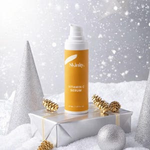 K&ouml;nnte beinhalten: Eine Vitamin-C-Serum-Flasche mit wei&szlig;er Pumpe und orangefarbenem Etikett, pr&auml;sentiert auf einer silbernen Geschenkbox mit goldenem Band. Auf dem Etikett steht "Skinity. Vitamin C Serum". Die Szene ist mit silbernen Ornamenten und Kunstschnee dekoriert.