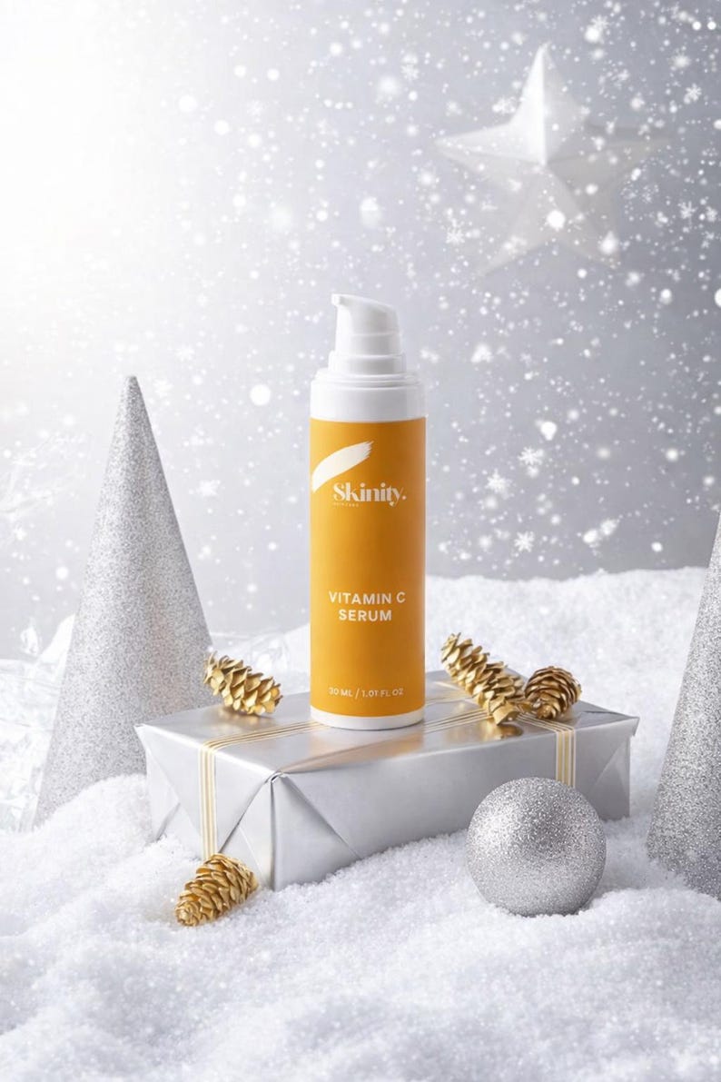 K&ouml;nnte beinhalten: Eine Vitamin-C-Serum-Flasche mit wei&szlig;er Pumpe und orangefarbenem Etikett, pr&auml;sentiert auf einer silbernen Geschenkbox mit goldenem Band. Auf dem Etikett steht "Skinity. Vitamin C Serum". Die Szene ist mit silbernen Ornamenten und Kunstschnee dekoriert.