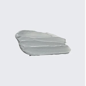 May include: A smear of light gray cosmetic product, possibly a mask or cream, against a white background. The product is thick and has visible texture from application, with several horizontal strokes. The overall appearance is smooth and opaque.